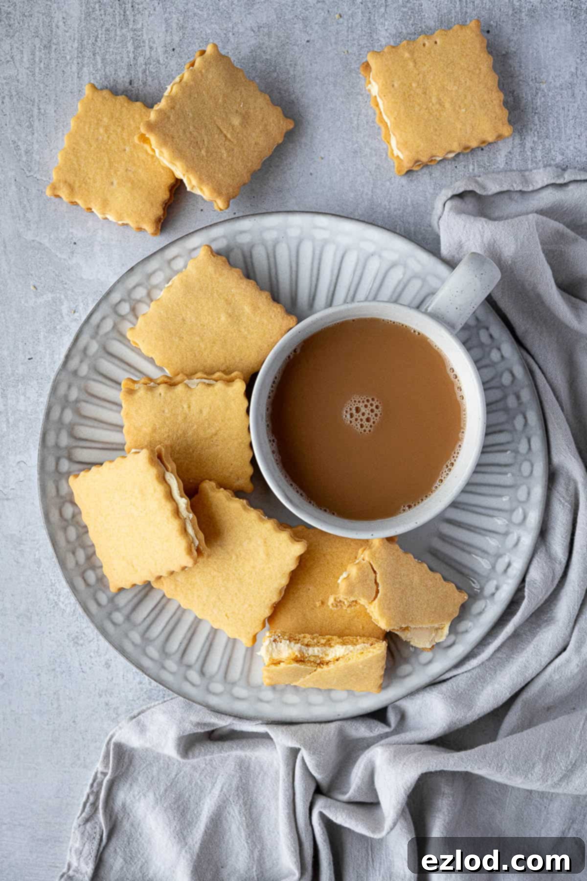 A pristine white plate holds several golden-yellow vegan custard creams, artfully arranged next to a comforting cup of tea, ready for enjoyment.