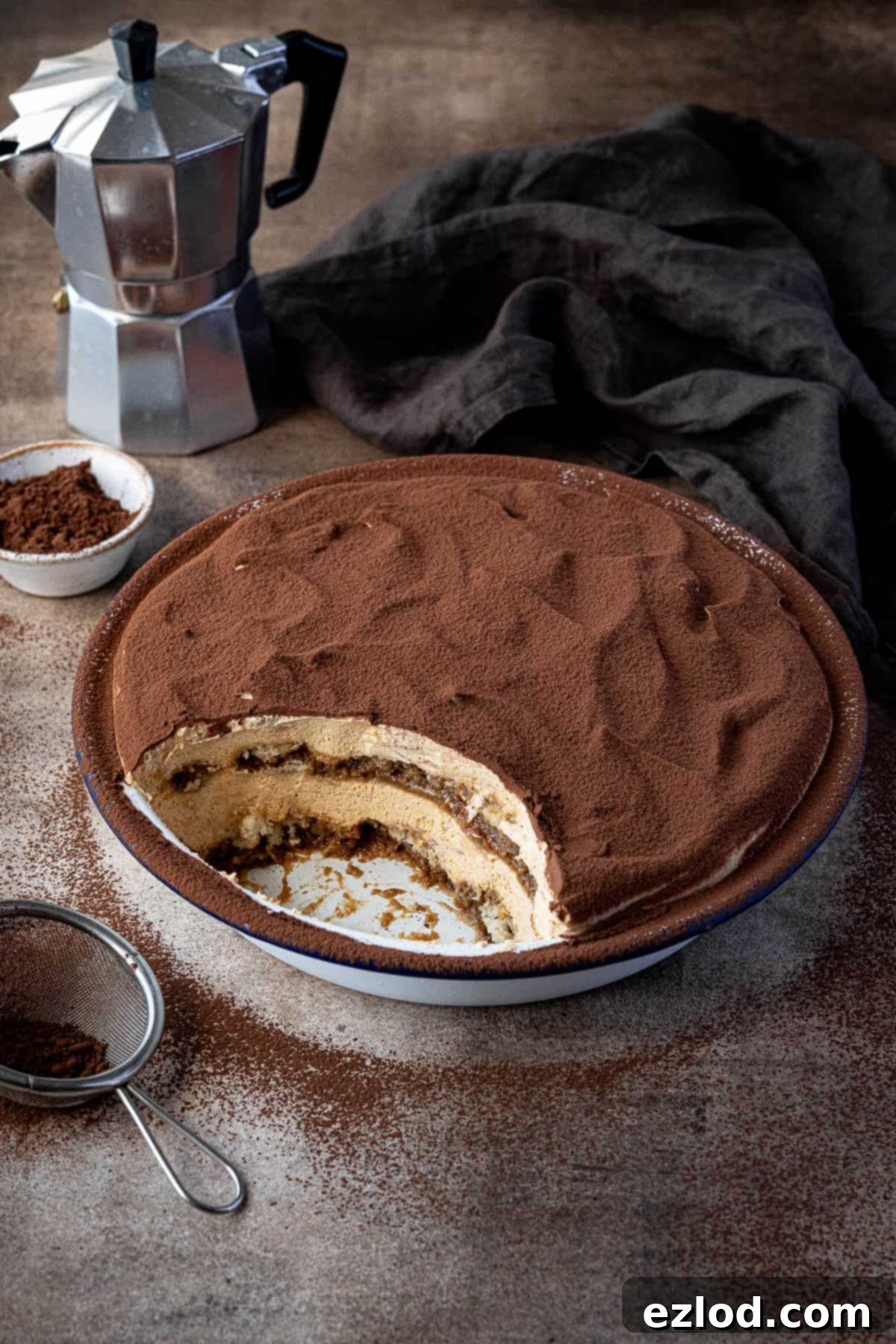 Pumpkin tiramisu in a round dish with a spoonful removed, a caffetiere, bowl of cocoa powder and small sieve in the background.