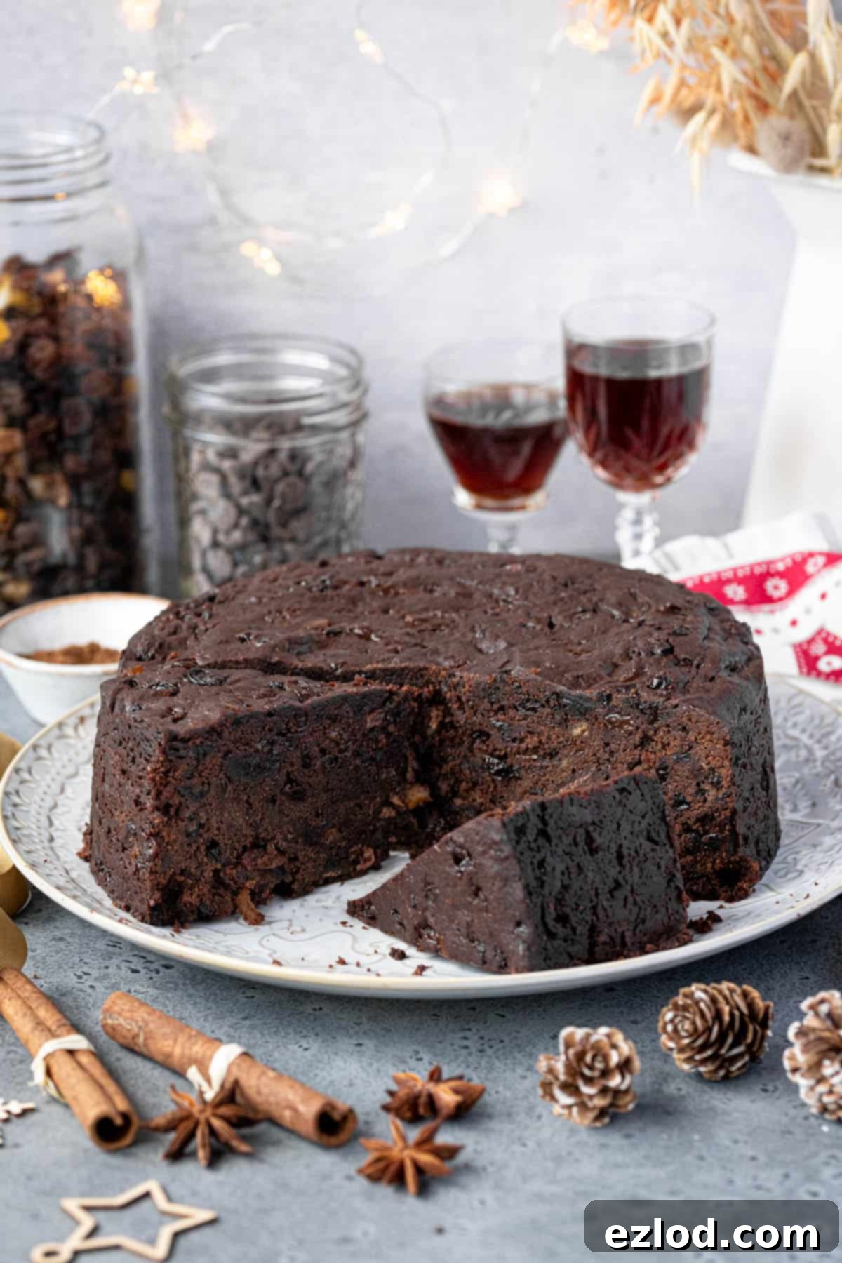 Sliced vegan chocolate Christmas cake on a white plate with glasses of rum, jars of dried fruit and chocolate chips and  whole spices.