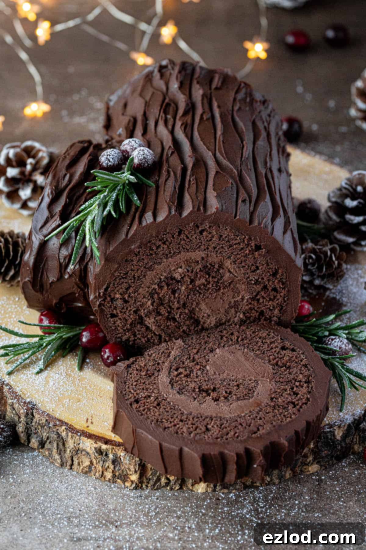 Plant-Based Chocolate Yule Log 2 A beautifully decorated vegan chocolate yule log with a slice removed, resting on a wooden board adorned with festive pine cones and red berries. It's the perfect Christmas dessert centerpiece.