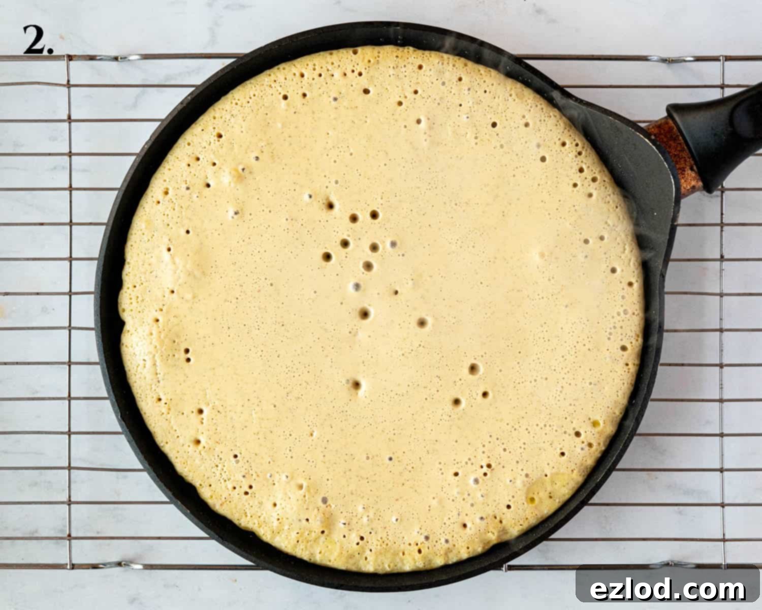 Golden Chickpea Fritters 6 Step 2 in the recipe: A savoury chickpea pancake cooking in a frying pan, showing bubbles forming on its surface and edges starting to set.