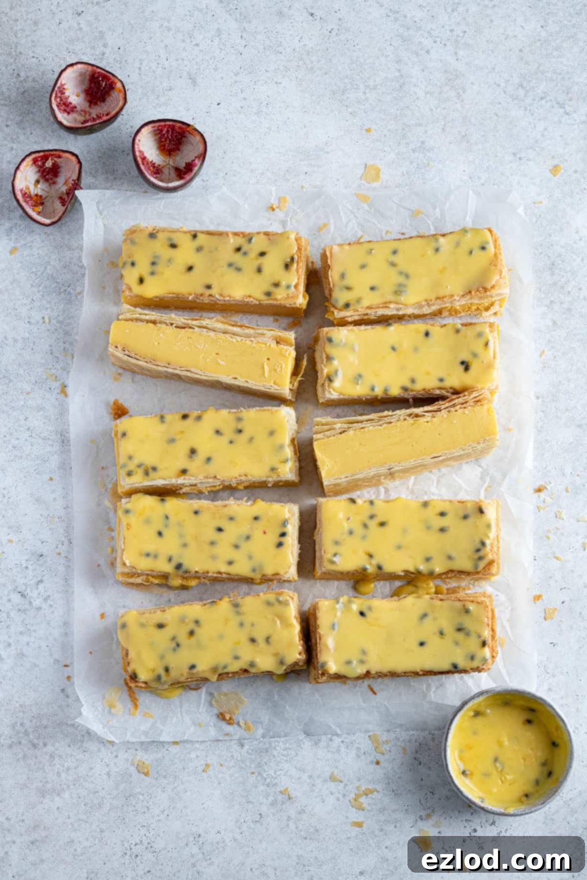 The sliced and glazed custard slices on a sheet of baking paper with passion fruit shells and a bowl of passion fruit glaze.