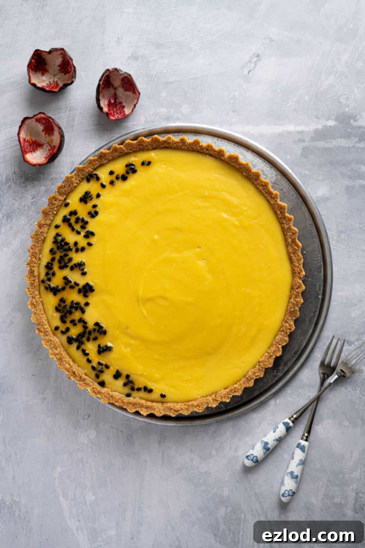 A stunning vegan passion fruit tart on a metal plate, ready to be served, accompanied by two forks and fresh passion fruit shells.