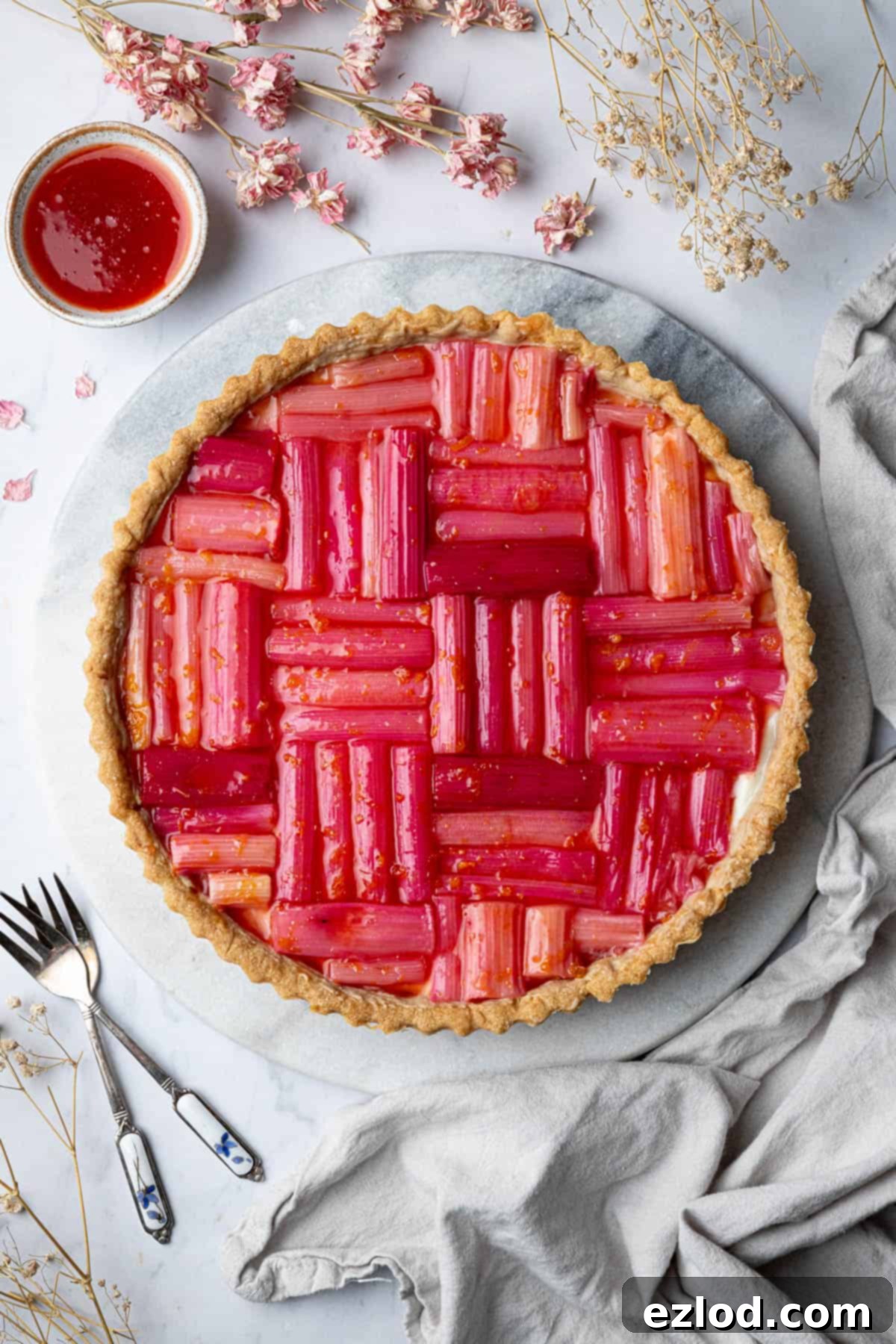 A stunning vegan rhubarb tart on a marble board, garnished with dried flowers, highlighting its crisp pastry, creamy filling, and vibrant pink roasted rhubarb.