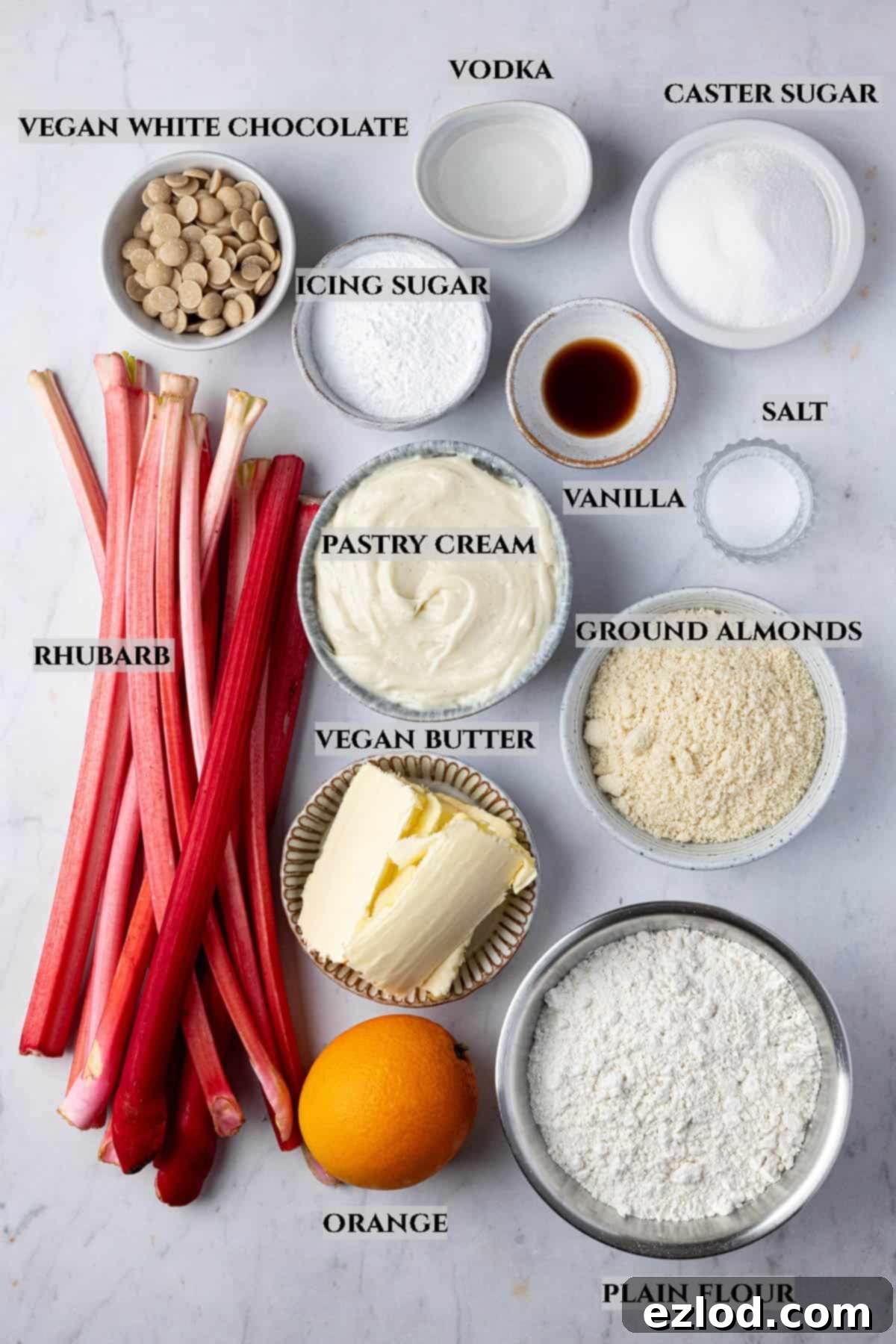 A collage of fresh ingredients laid out on a surface, clearly labelled, for making a vegan rhubarb tart including rhubarb stalks, oranges, vegan butter, flour, sugar, and vanilla extract.