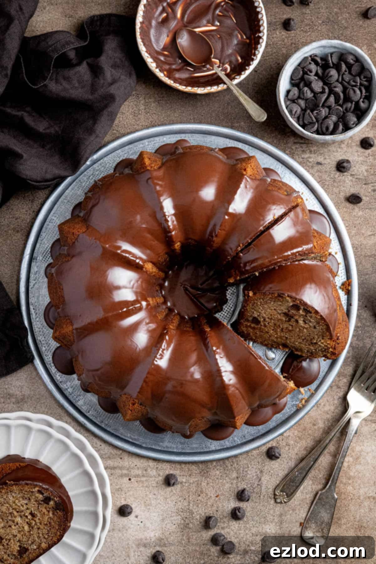 Vegan banana bundt cake on a metal plate with a couple of slices removed surrounded by bowls of chocolate chips and chocolate glaze.