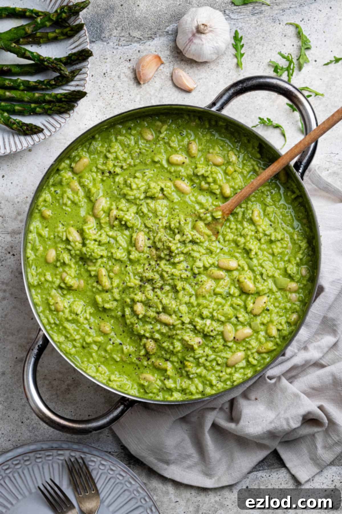 The cooked rocket pesto risotto presented in a pan surrounded by serving plates, forks, fresh garlic, and a side of grilled asparagus.
