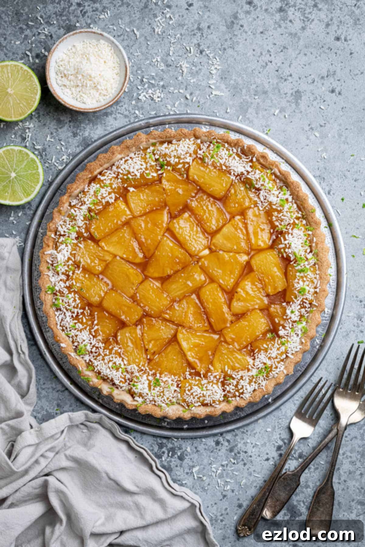A beautifully presented Caramelised Pineapple Coconut Tart on a stylish metal plate, garnished with fresh lime slices and a small bowl of desiccated coconut in the background, inviting forks poised for a taste.