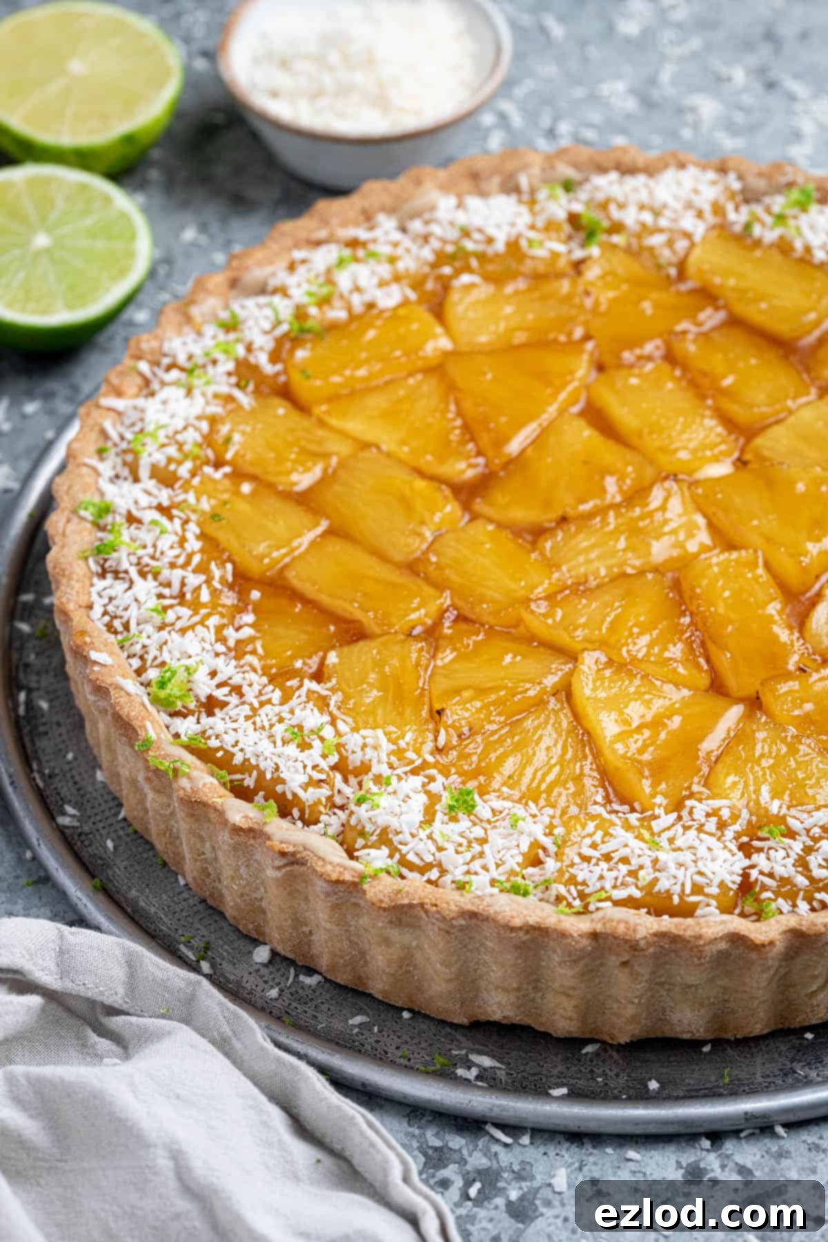 A close-up view of the golden-brown caramelised pineapple topping on the coconut tart, with blurred lime slices and a bowl of desiccated coconut adding to the tropical ambiance in the background.