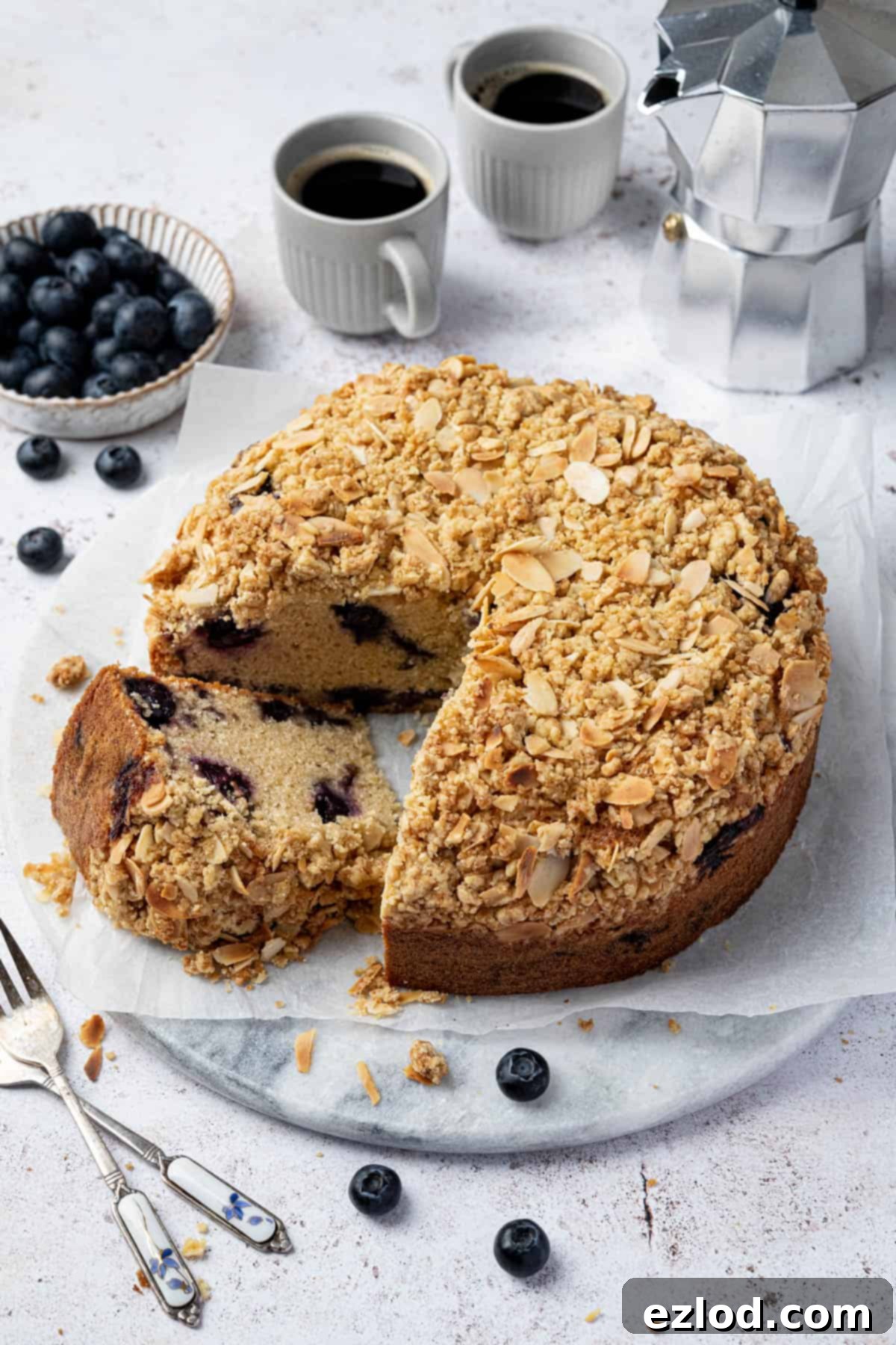 A beautifully sliced vegan blueberry crumb cake on a marble board, accompanied by fresh blueberries and coffee cups in the background.