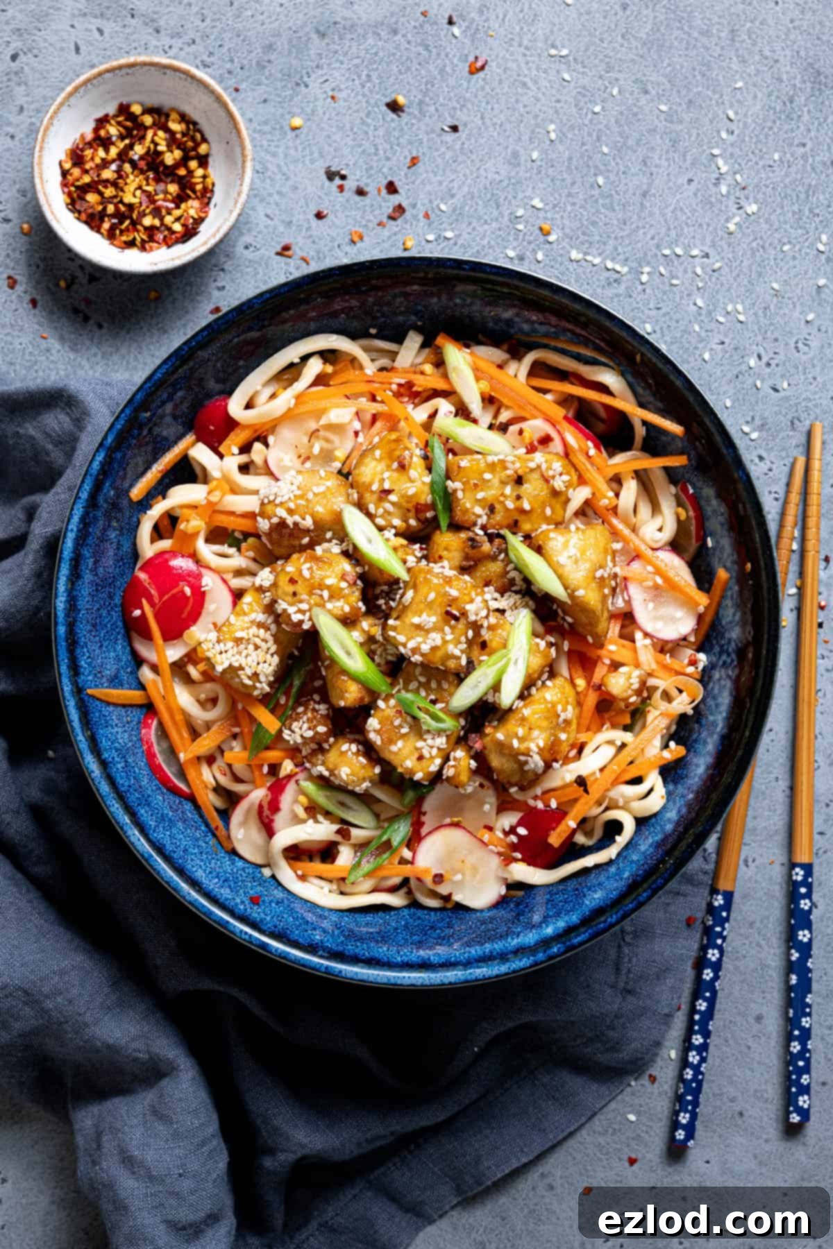 Sticky tofu noodle salad in a blue bowl with chopsticks and a bowl of chilli flakes.