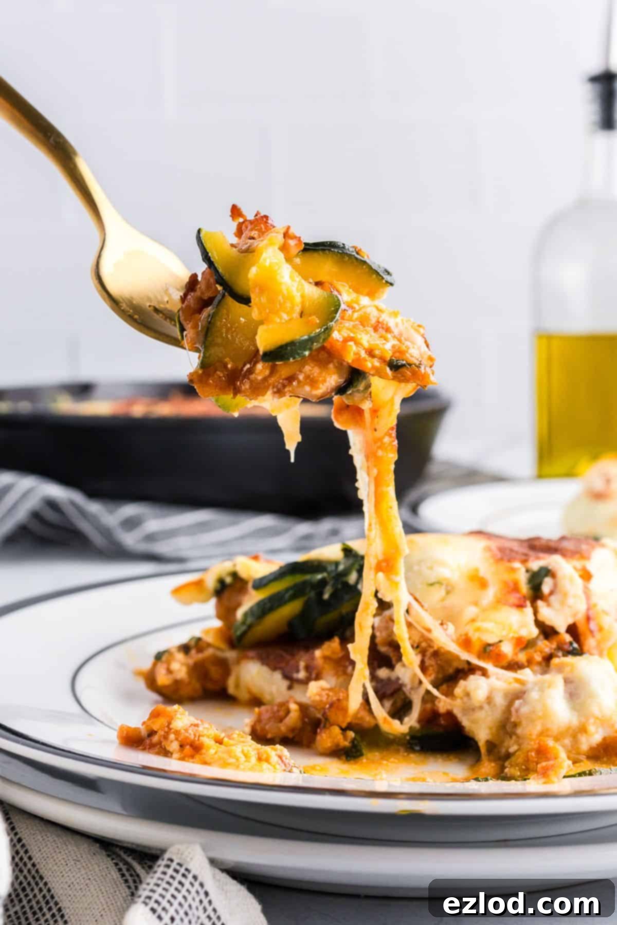 High Protein One Pan Zucchini Lasagna 3 A close-up shot of a fork lifting a perfect bite of zucchini lasagna, showing distinct layers of tender zucchini, melted golden cheese, savory Italian sausage, and rich marinara sauce. In the background, the rest of the bubbling, freshly baked lasagna sits in a cast iron pan, ready to be enjoyed.