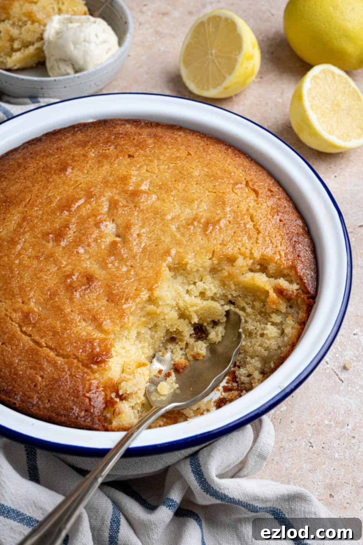 A close-up shot of the baked lemon elderflower almond pudding in its dish, with a spoonful removed to show its moist texture, and elegant lemon slices in the blurred background.