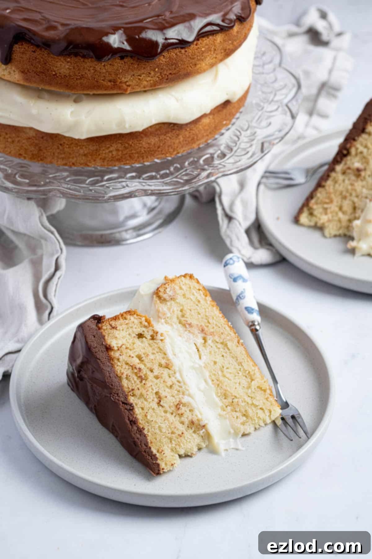 A beautifully presented slice of vegan Boston cream pie on a grey plate with a fork, showcasing the fluffy vanilla cake, creamy pastry filling, and glistening chocolate ganache.