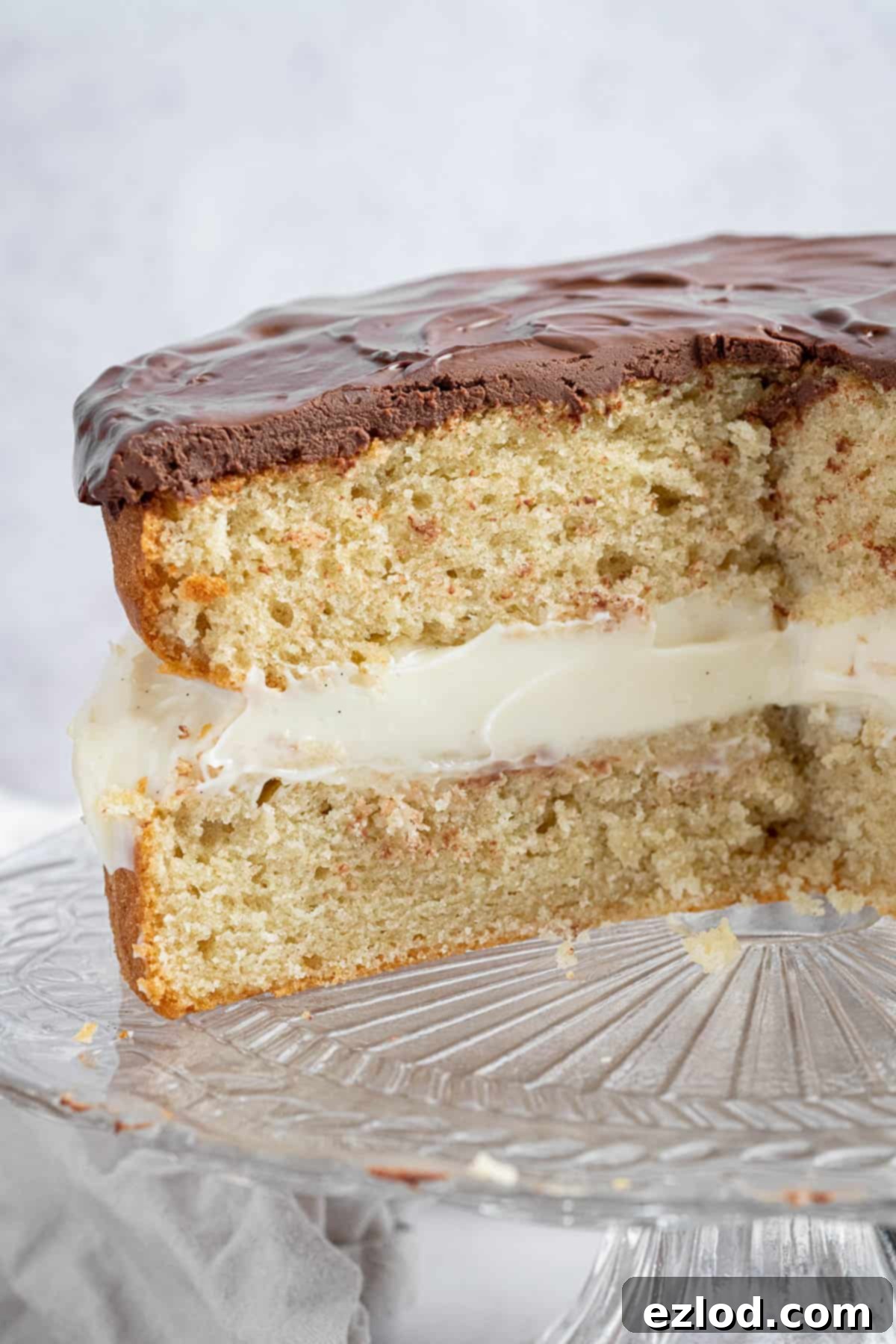 A close-up view of a sliced vegan Boston cream pie, revealing the exquisite layers of light vanilla cake, creamy vanilla pastry cream, and rich chocolate ganache.