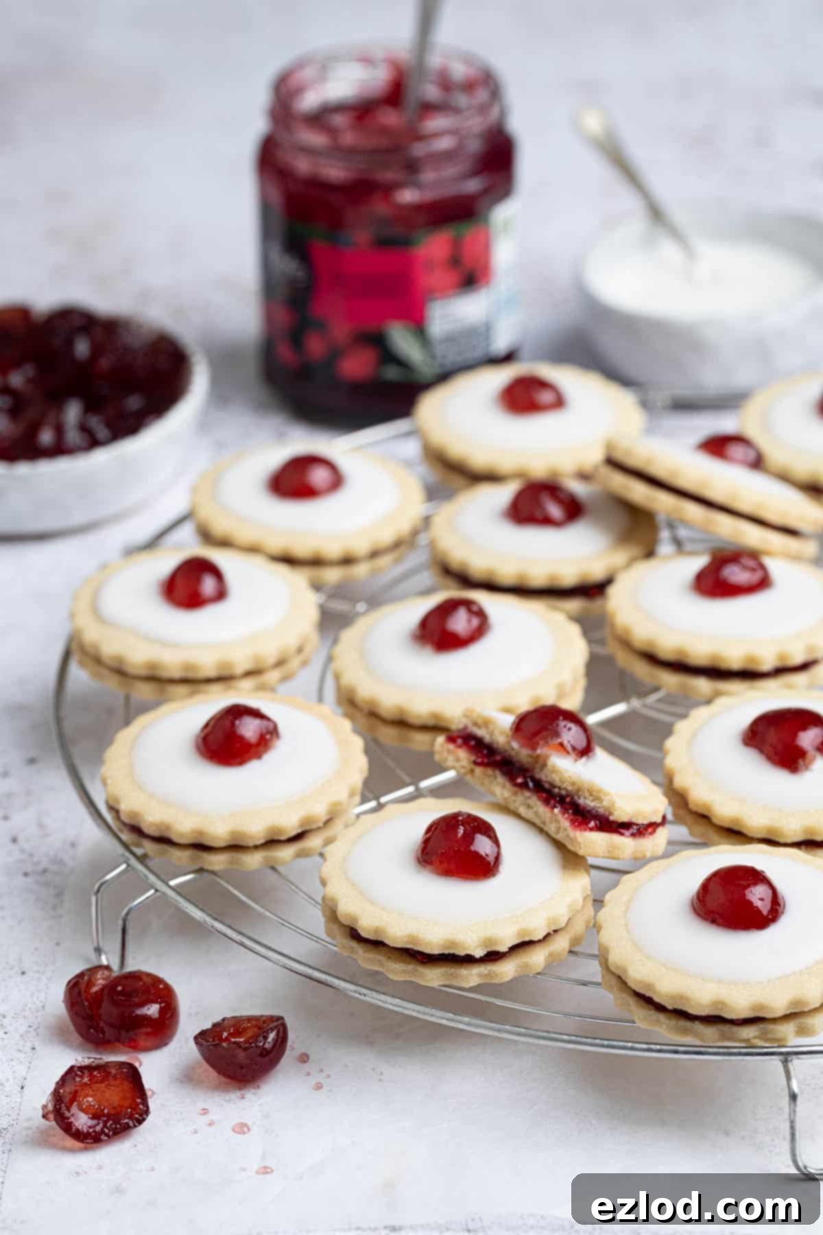 Vegan empire biscuits on a round wire cooling rack with a jar of jam and bowls of glace cherries and icing in the background.