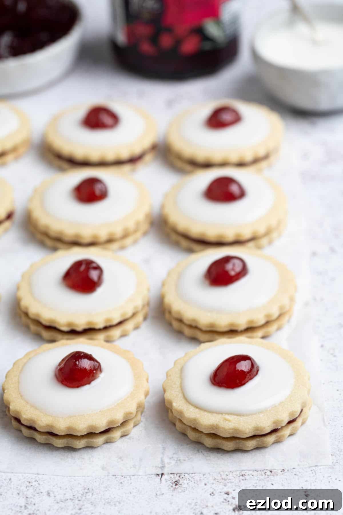 Two rows of empire biscuits with a jar of jam and bowls of glace cherries and icing in the background.