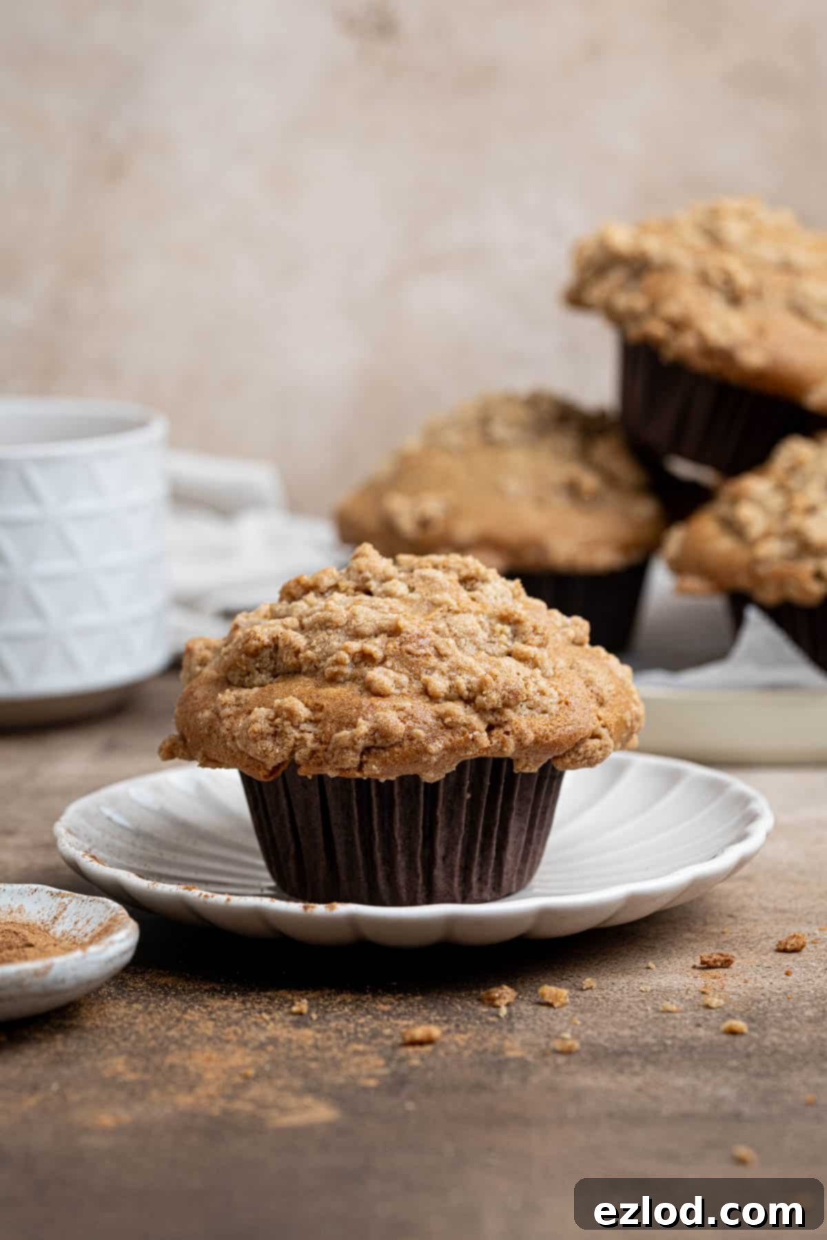Spiced Pear Plant-Based Muffins 2 A beautifully baked vegan cinnamon pear muffin on a white plate, with a stack of more muffins and a cup of steaming tea blurred in the background, highlighting its tall dome and streusel topping.