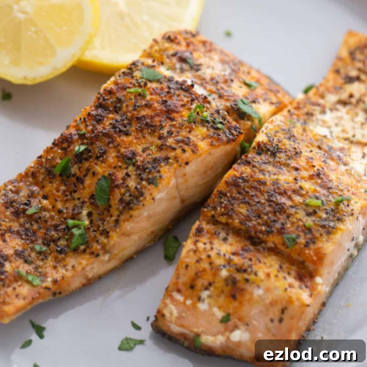 A plate of two air fryer salmon fillets seasoned with lemon pepper seasoning and garnished with fresh herbs and lemon. 