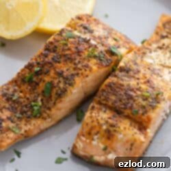 A white plate with seasoned air fryer salmon garnished with two slices of lemon on the side.