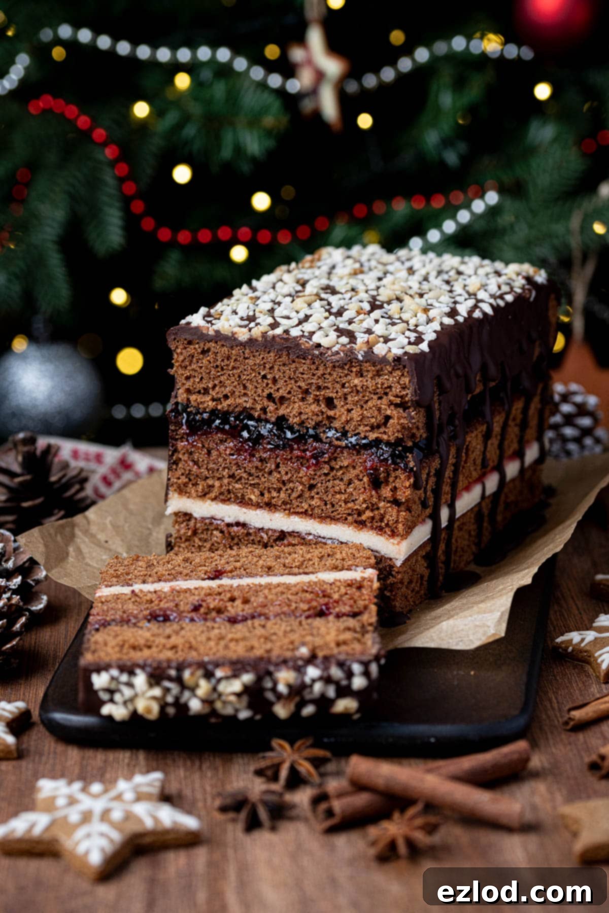 A beautifully garnished Vegan Piernik cake, glistening with a chocolate glaze and sprinkled with chopped nuts, with one slice elegantly cut, displayed on a metal tray in front of a festive Christmas tree.