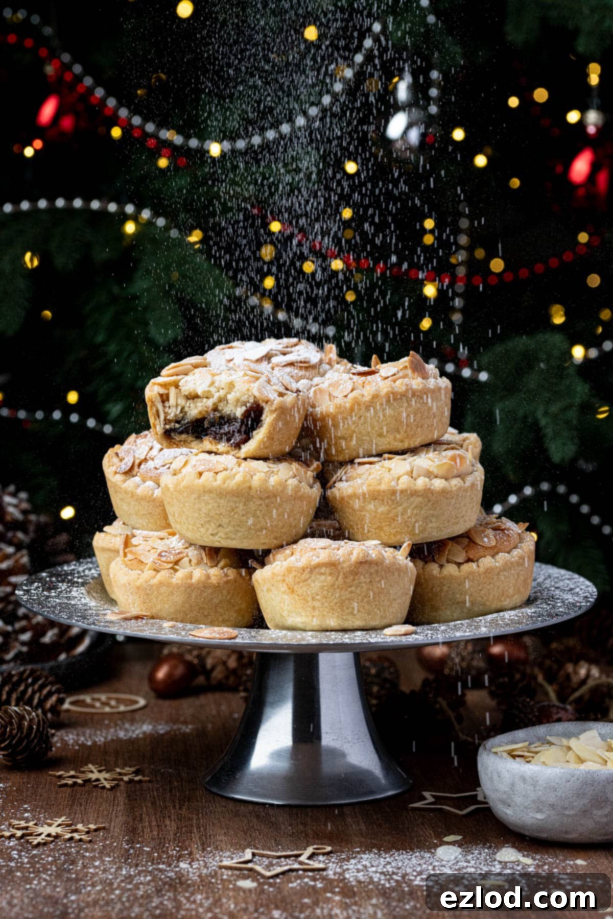 A stack of vegan frangipane mince pies on a cake stand being dusted with icing sugar in front of a Christmas tree.