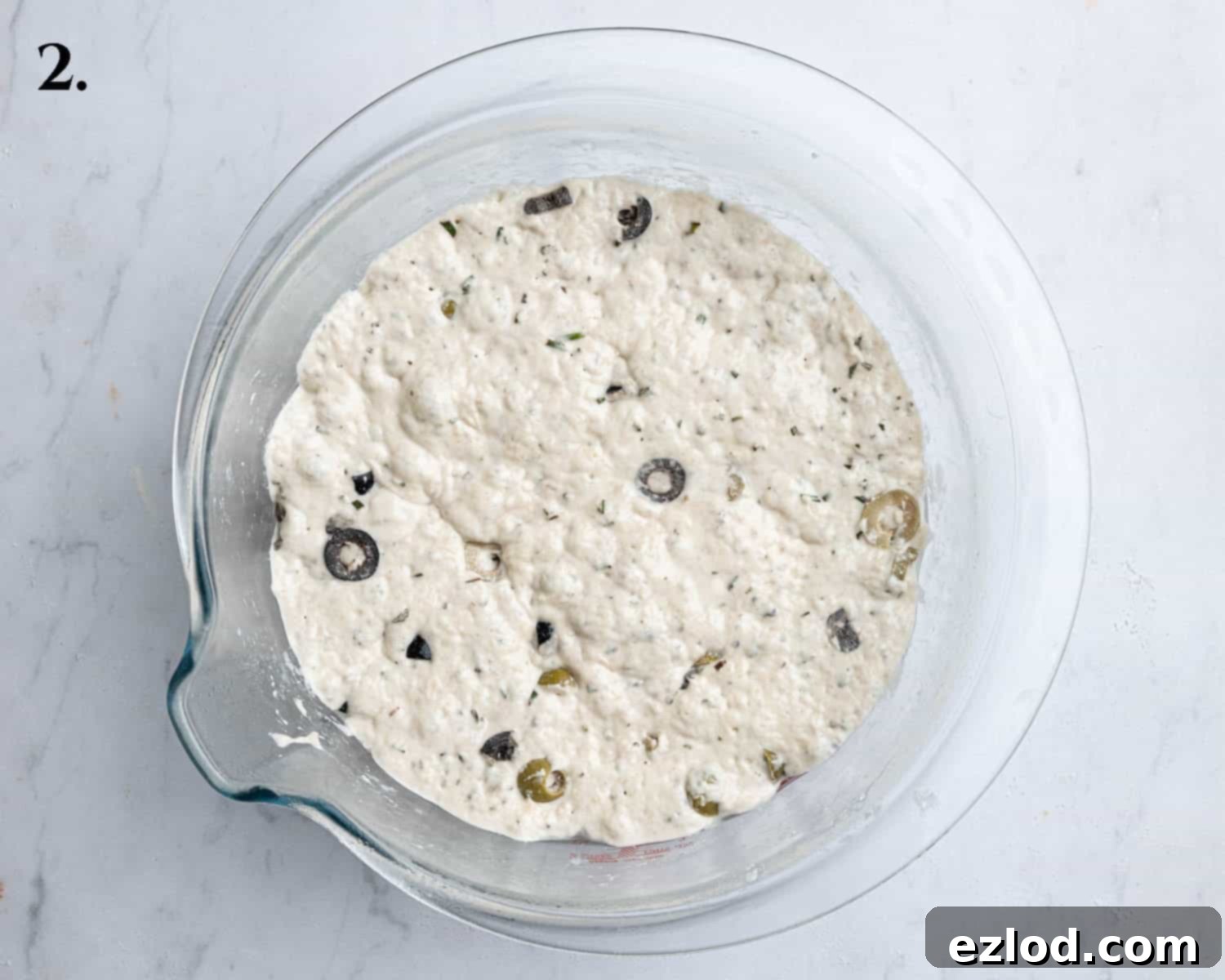 Step 2 of no-knead olive bread: The dough, now significantly risen and bubbly after its initial and overnight fermentation, rests in a bowl, visibly expanded and light.