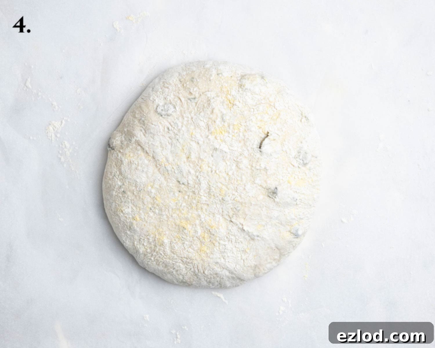Step 4 of the no-knead olive bread recipe: The partially shaped dough now sits on a square of baking paper, ready for its final rise, with a light dusting of flour on top.