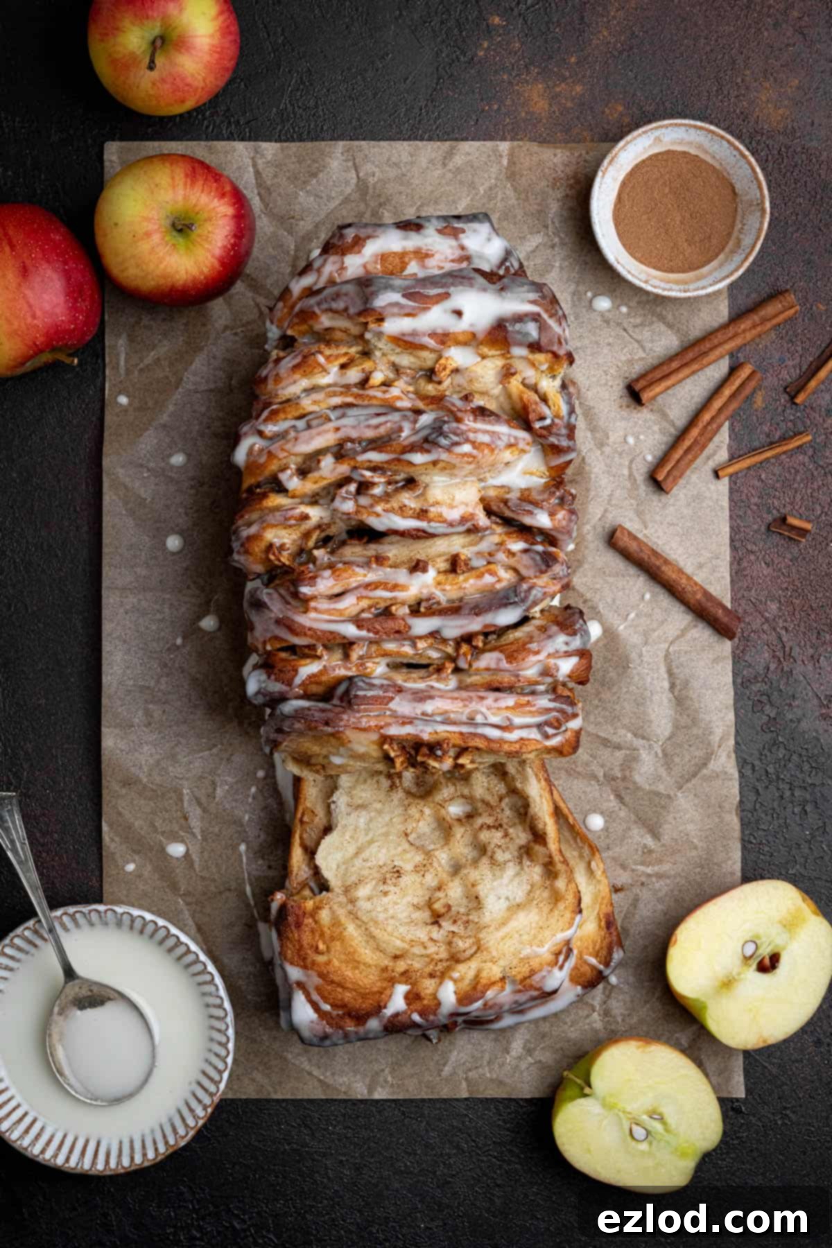 A beautifully baked vegan apple cinnamon pull apart bread loaf on a sheet of baking paper, surrounded by fresh apples, cinnamon sticks, and a bowl of vanilla glaze.