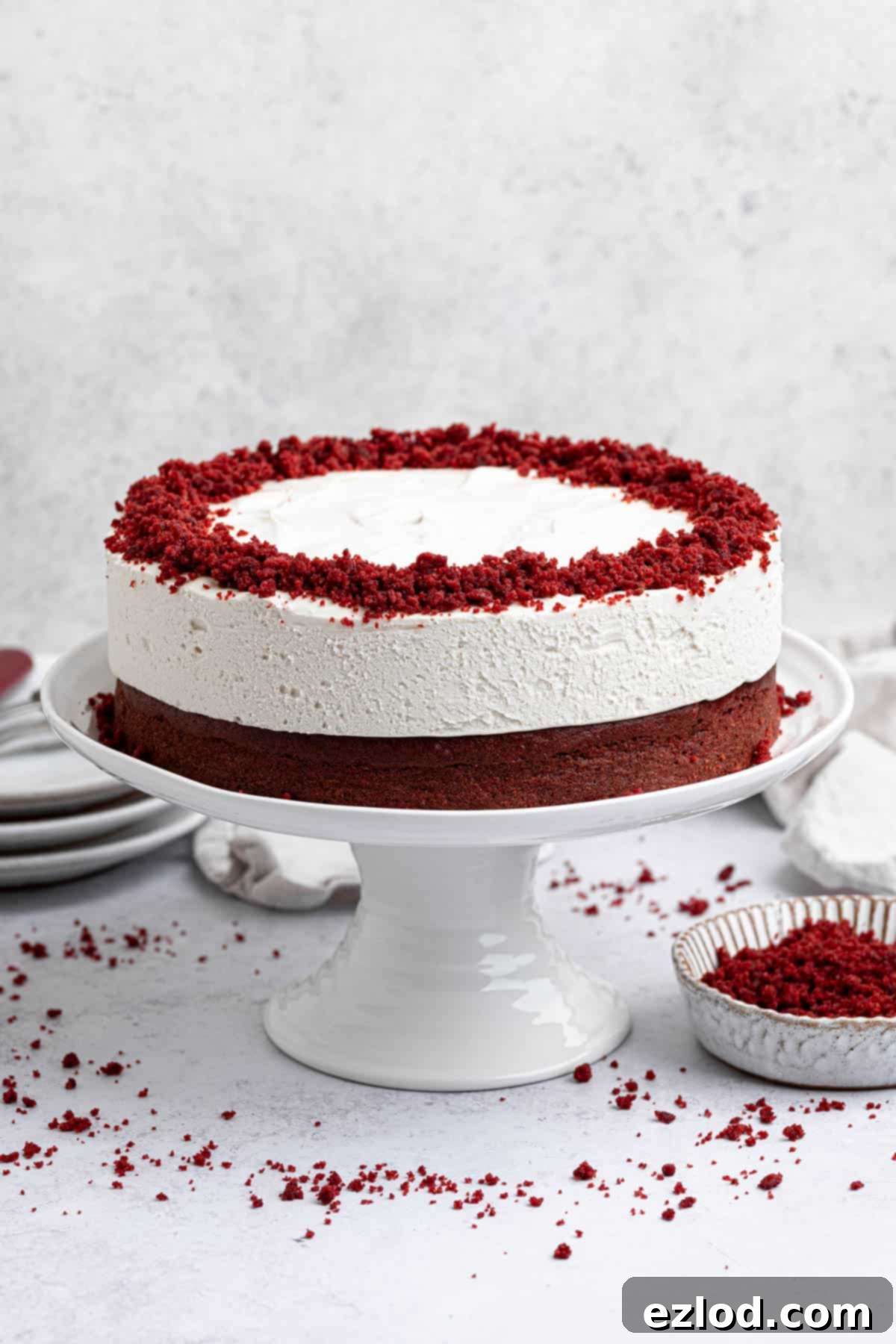 Red velvet cheesecake cake on a white cake stand with a bowl of red velvet cake crumbs.
