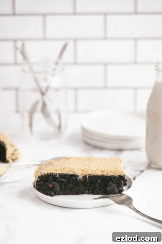 Side view of a generous slice of Gluten-Free Chocolate Cake with creamy Peanut Butter Frosting, showcasing the beautiful layers on a white rimmed plate. A fork is ready for a bite, with a glass of milk and extra plates blurred in the background, suggesting a delightful gathering.
