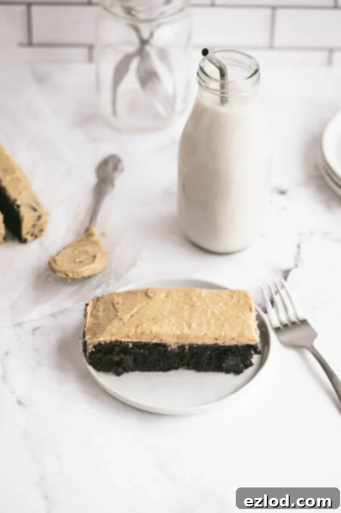 A close-up, slightly angled overhead shot of a slice of Healthy Gluten-Free Chocolate Cake with generous Peanut Butter Frosting on a white plate. A glass of milk and a fork are positioned next to it, evoking a sense of imminent enjoyment and healthy indulgence.