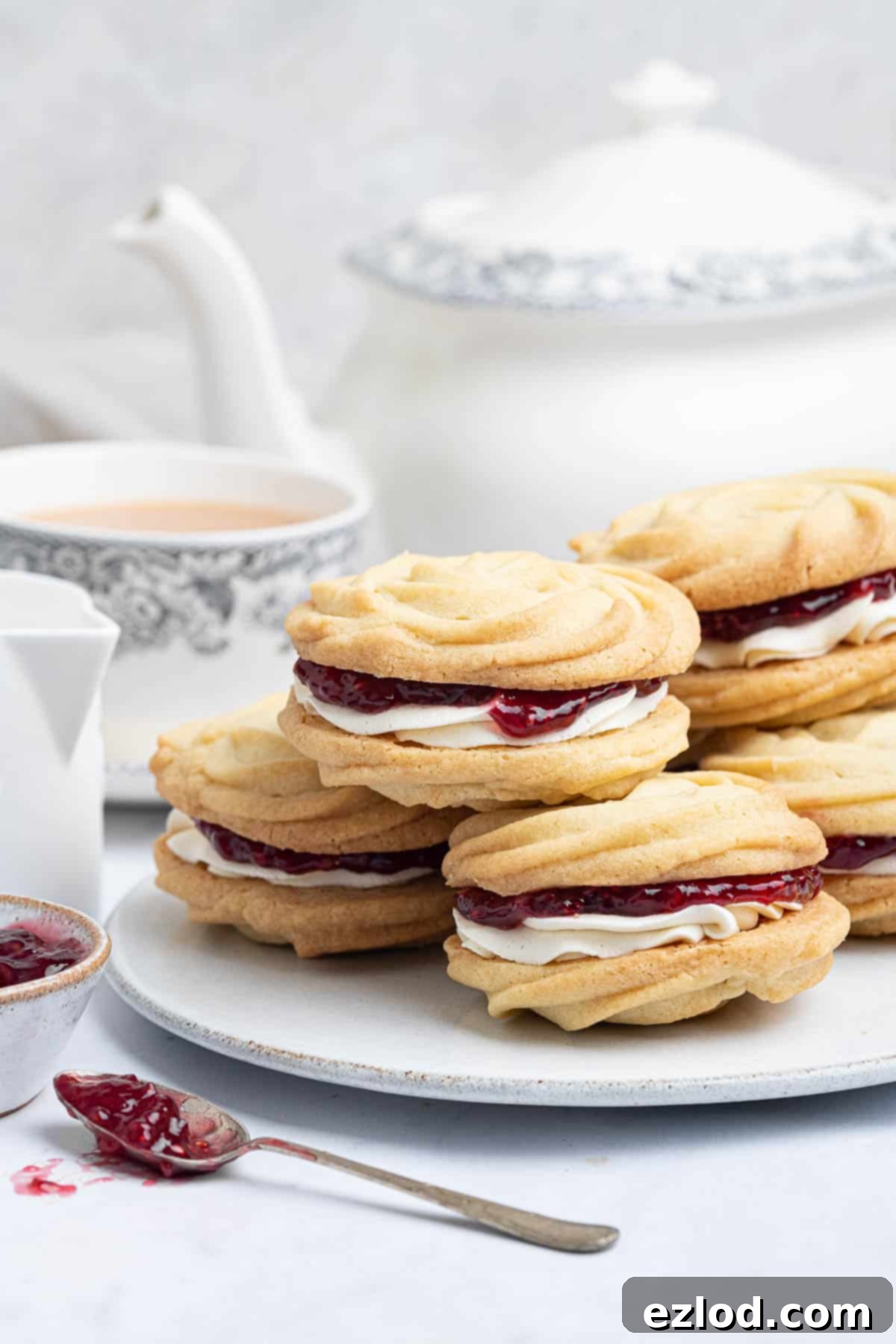Dairy-Free Viennese Delights 4 Vegan Viennese whirls elegantly arranged on a plate, accompanied by a teapot, a cup of tea, and a small bowl of jam, setting the scene for afternoon tea.