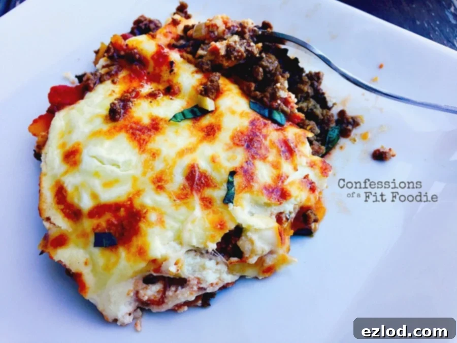 Close up photo of lazy butternut squash lasagna on a white plate with a fork in the meal, ready to dig in. Ground beef and tomato sauce peek out from a layer of crispy lasagna cheese and garnished with some basil.