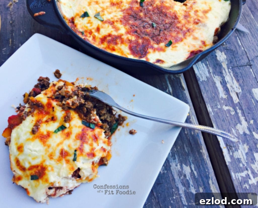 Photo of a serving of lazy butternut squash lasagna on a white plate with a fork ready to dig in. Ground beef and tomato sauce peek out from a layer of crispy lasagna cheese and garnished with some basil. In the background is the rest of the lasagna in a cast iron skillet sitting on a wooden table.