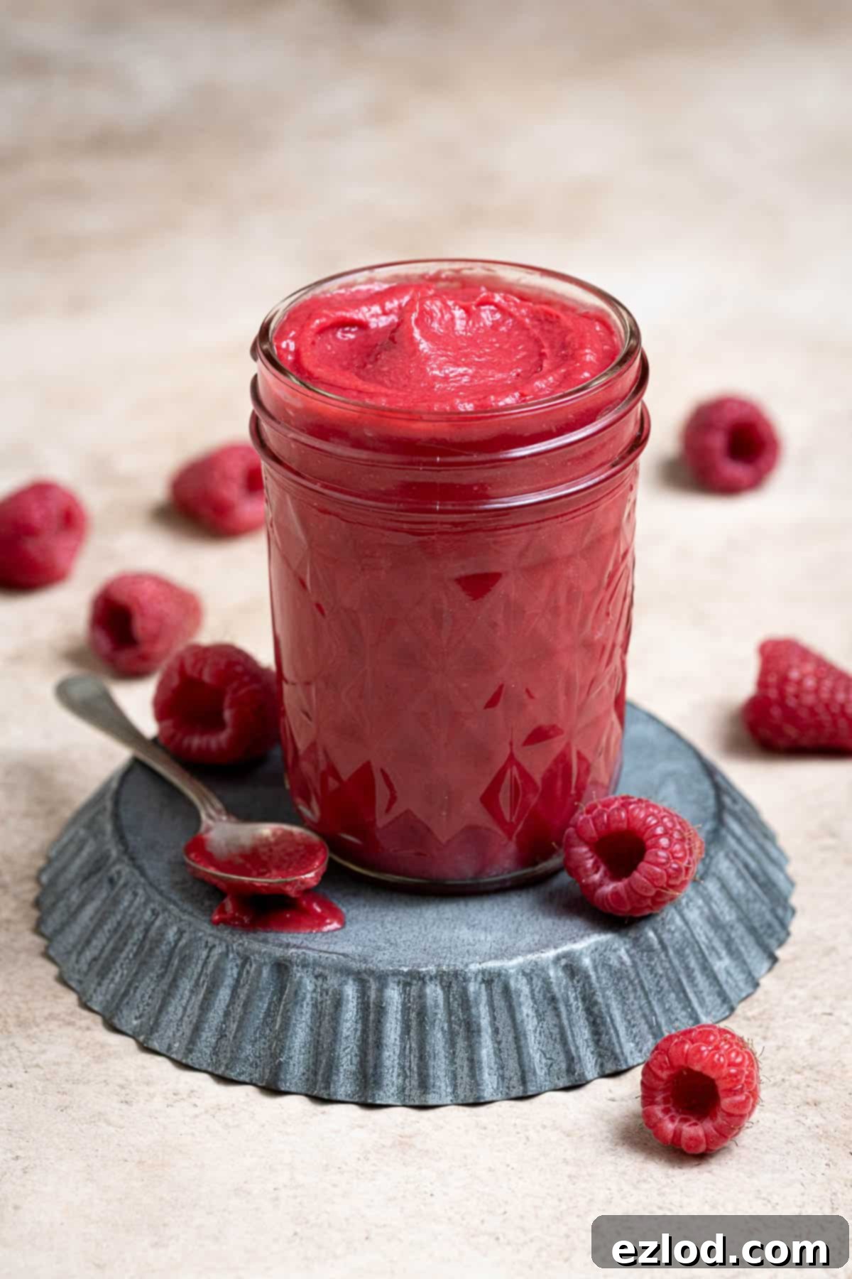 A glass jar filled with vibrant vegan raspberry curd, garnished with fresh raspberries and a spoon, resting on a rustic metal tray.
