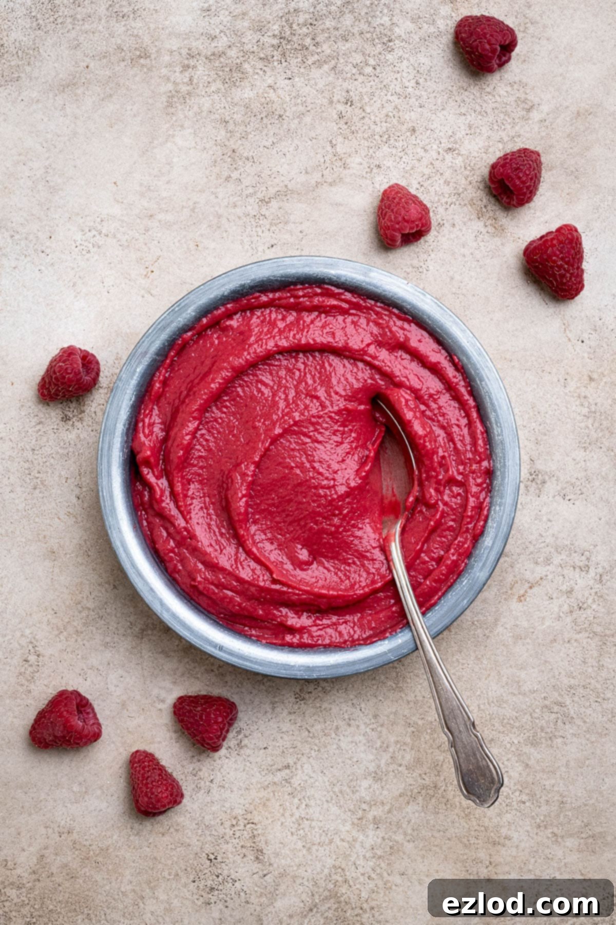 A charming bowl of smooth vegan raspberry curd, accompanied by a spoon and scattered fresh raspberries, ready to be enjoyed.