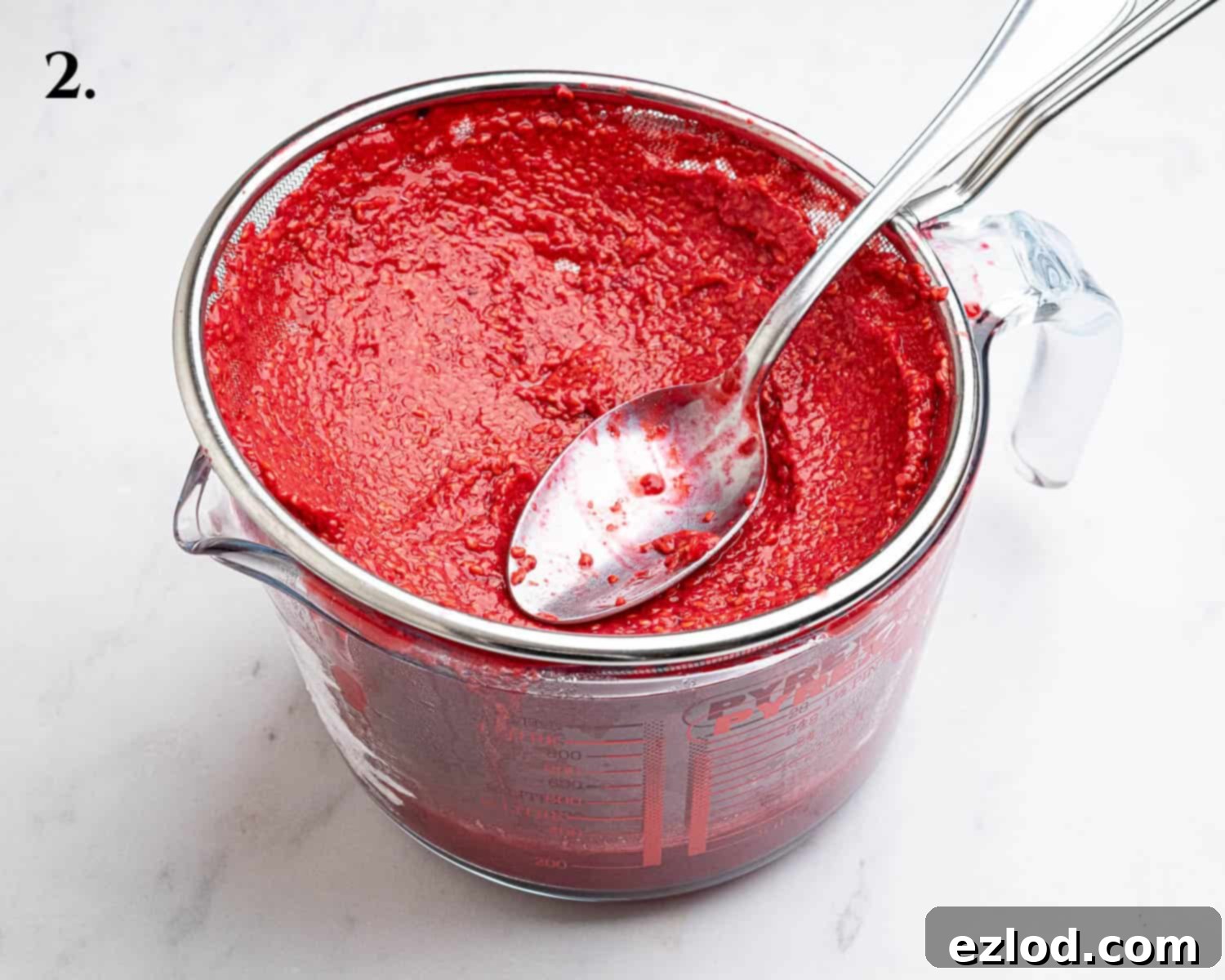 Step 2 in making vegan raspberry curd: A close-up of raspberry puree being pressed through a fine-mesh sieve with a spatula to remove seeds.