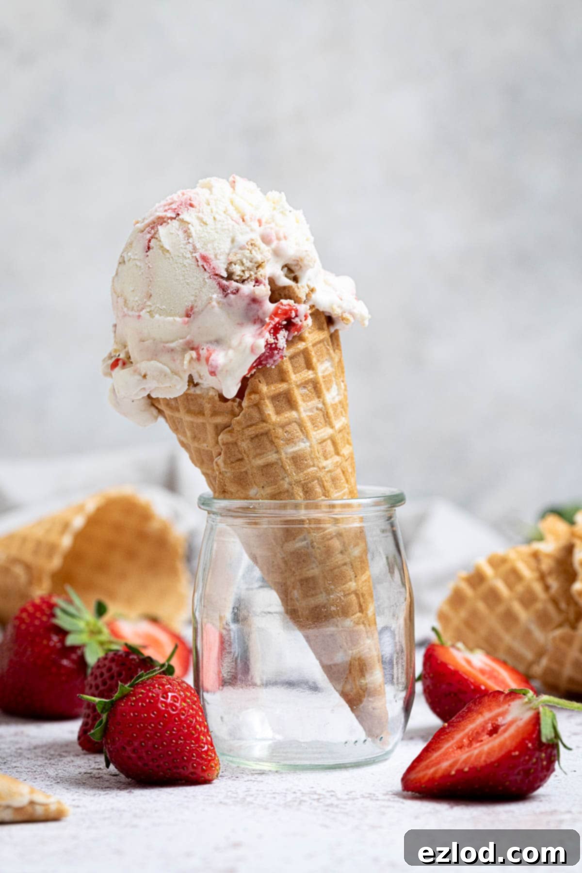 A scoop of homemade vegan strawberry cheesecake ice cream in a glass jar, surrounded by fresh strawberries and waffle cones.