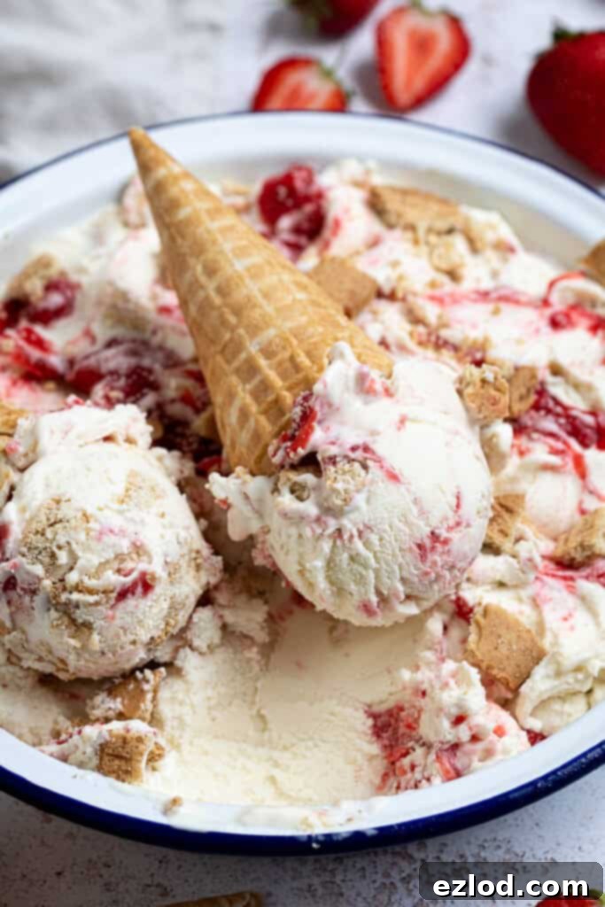 Two generous scoops of vegan strawberry cheesecake ice cream sitting atop a container filled with more ice cream, with fresh strawberries around.
