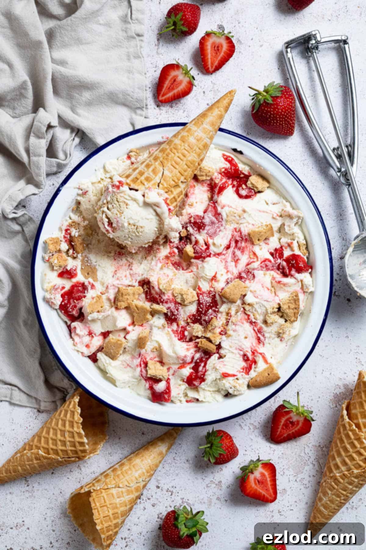 A container full of vegan strawberry cheesecake ice cream with an ice cream cone on top, surrounded by fresh strawberries, empty ice cream cones, and an ice cream scoop.