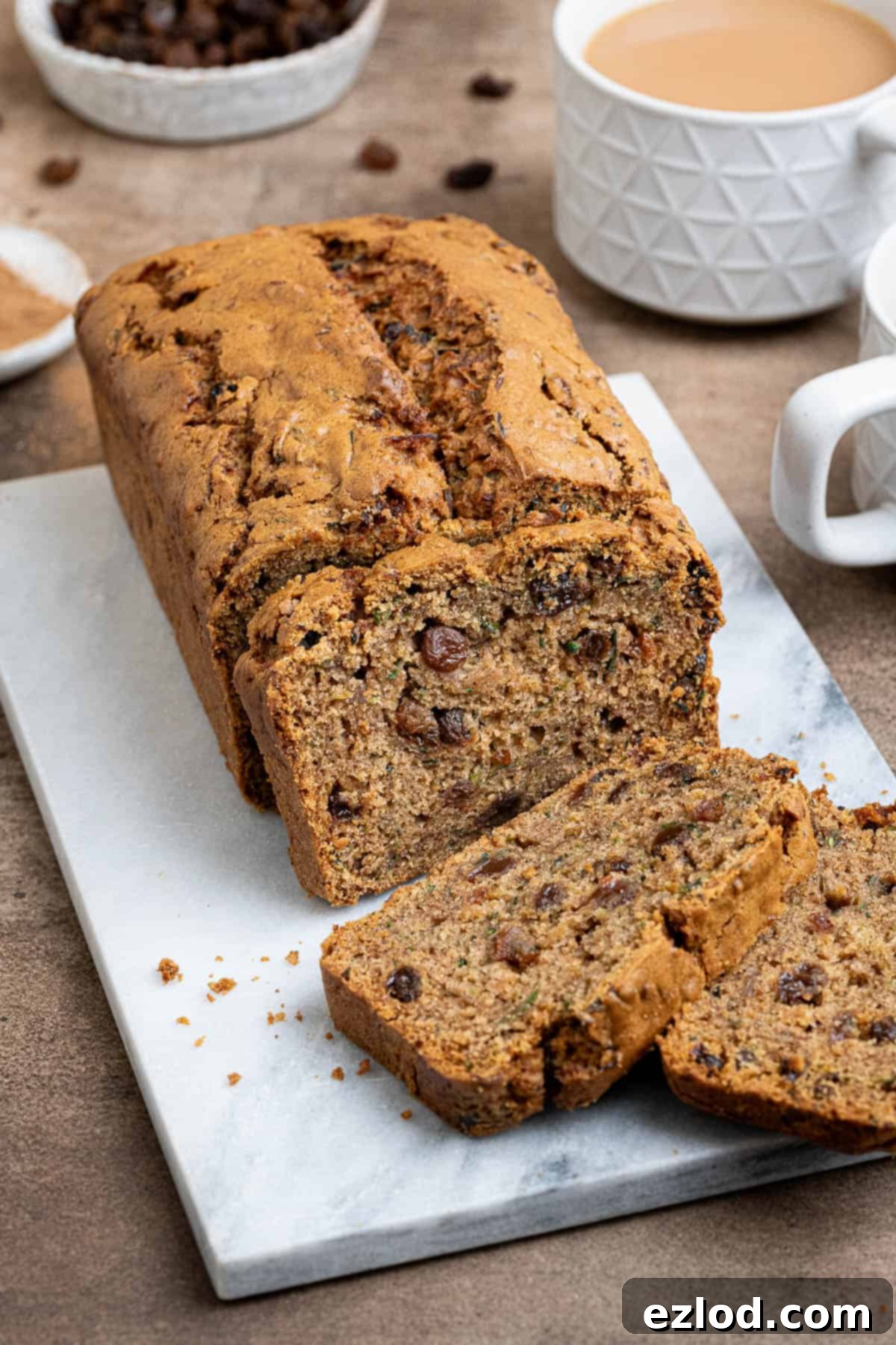 Vegan Zucchini Spice Cake 2 Vegan spiced courgette cake on a marble board with cups of tea and bowls of sultanas and cinnamon, showcasing its moist texture and rich color.