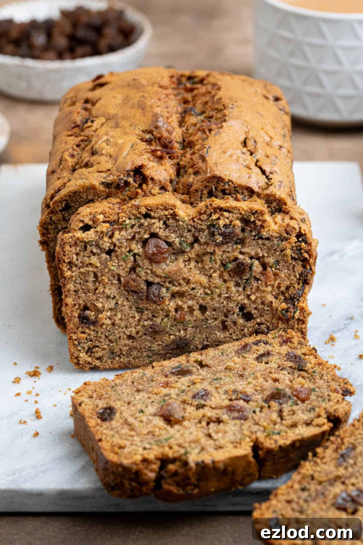Vegan Zucchini Spice Cake 4 Sliced vegan courgette cake on a marble board with a cup of tea and a bowl of sultanas, showing off the perfectly baked interior and delicious crumb.