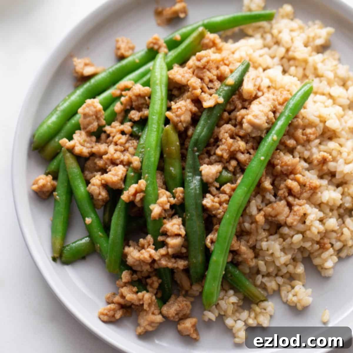 A plate of ground chicken stir fry on a bed of brown rice and stopped with steamed green beans.