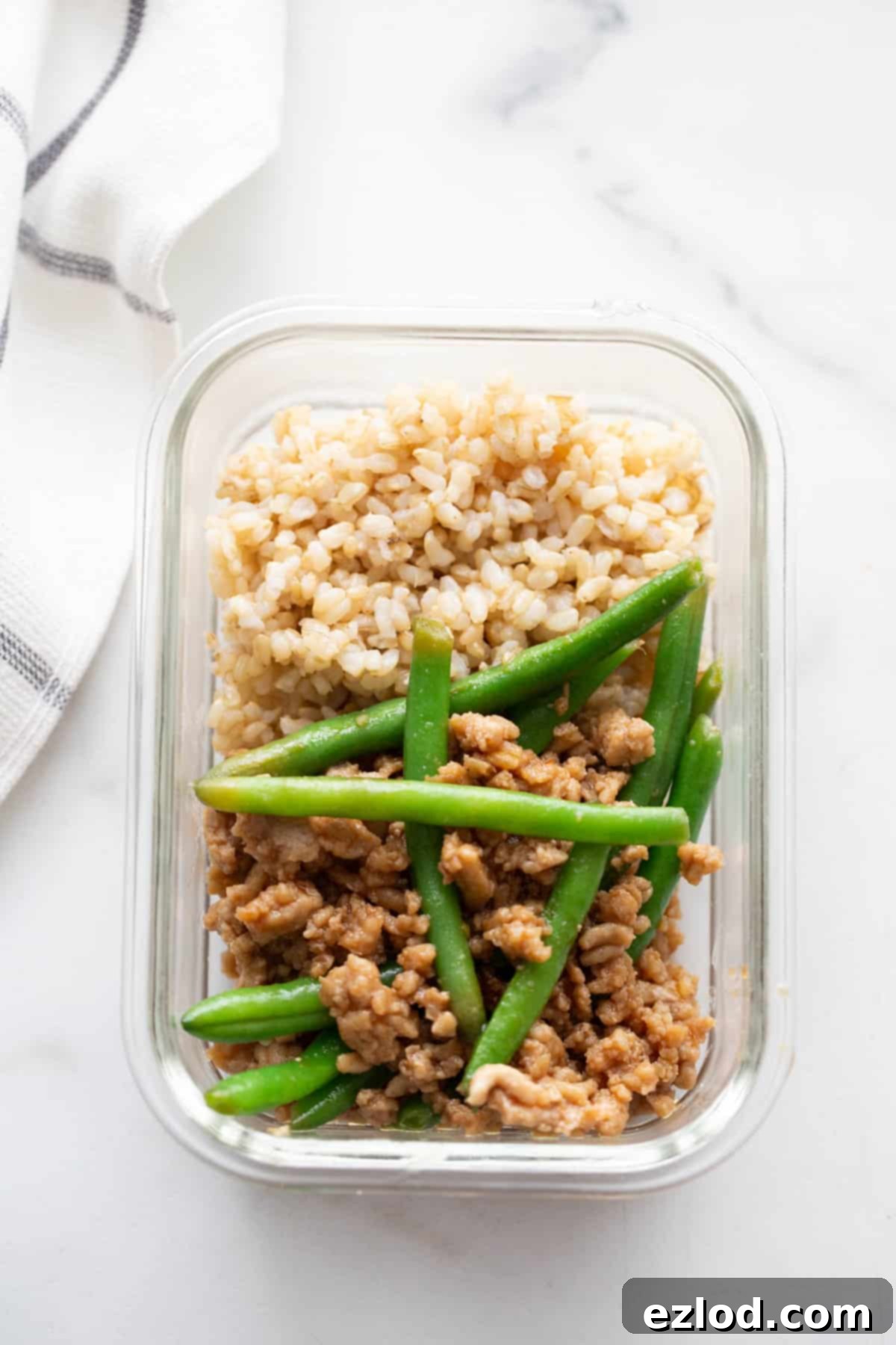 A glass meal prep container with brown rice and ground chicken stir fry and green beans.
