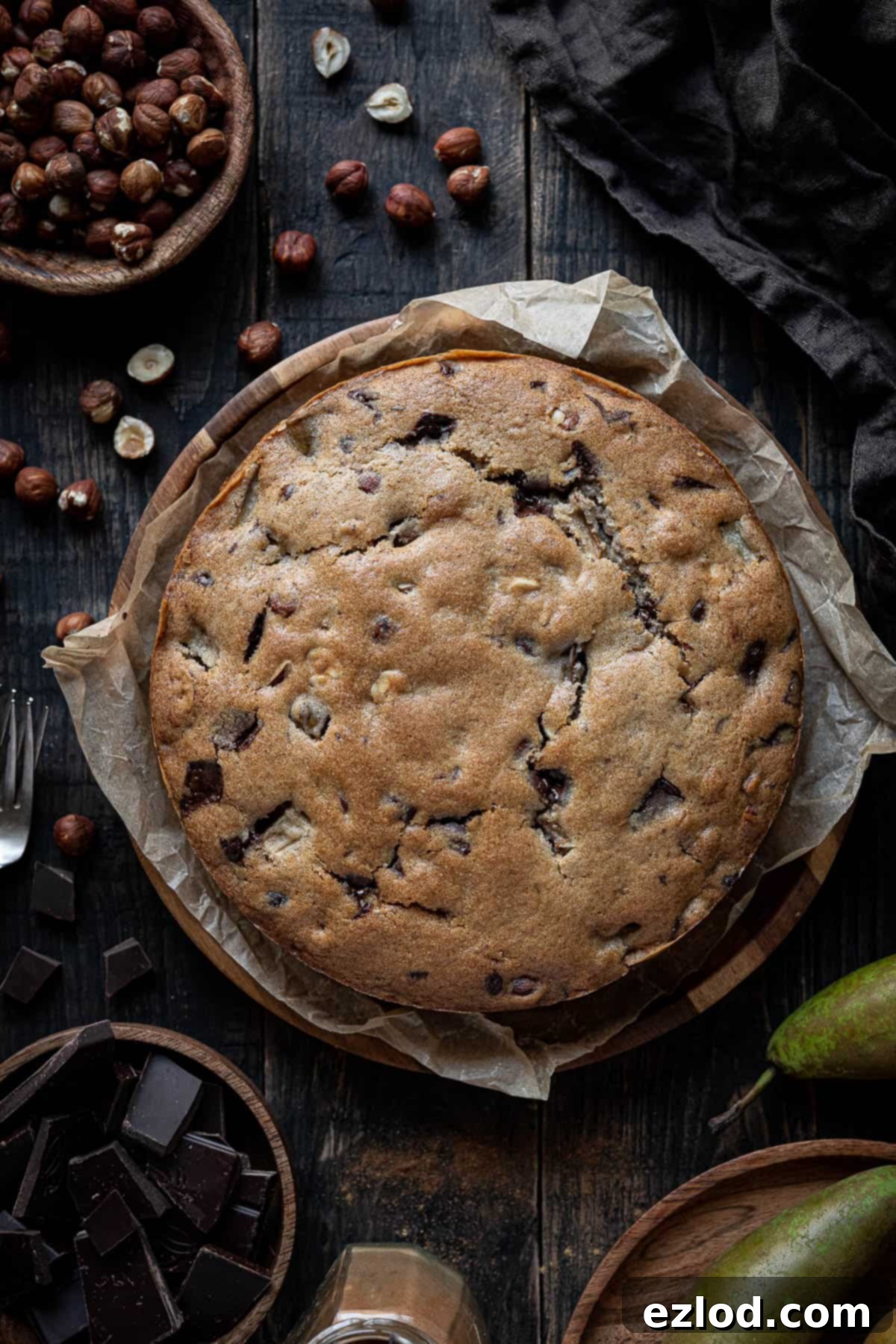 A beautifully sliced vegan chocolate, pear, and hazelnut cake on a wooden plate, surrounded by bowls of fresh pears, dark chocolate chunks, and whole hazelnuts, set on a rustic wooden table.