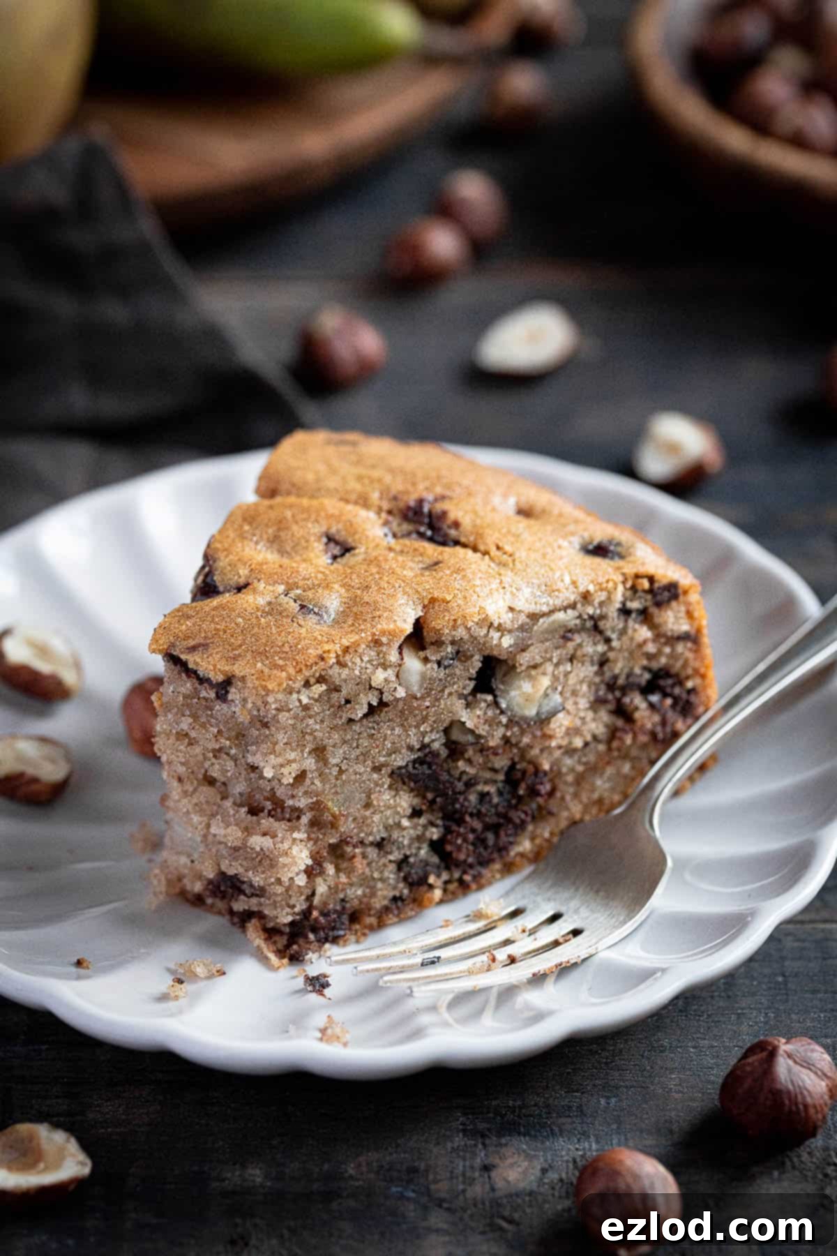 A close-up view of a single slice of vegan chocolate, pear, and hazelnut cake, showcasing the moist texture, visible chocolate chunks, and pear pieces within the golden-brown crumb.