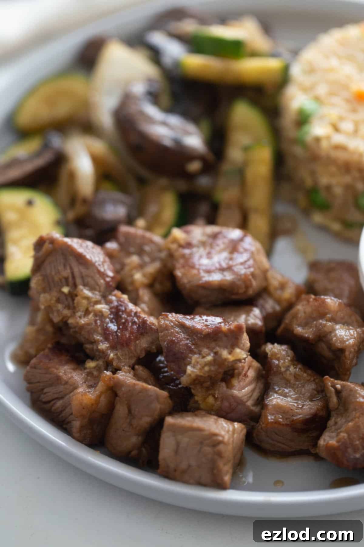 Sizzling Hibachi Steak 3 Cubed steak drizzled with ginger sauce on a plate; sauteed veggies and rice are on the same plate in the background, out of focus