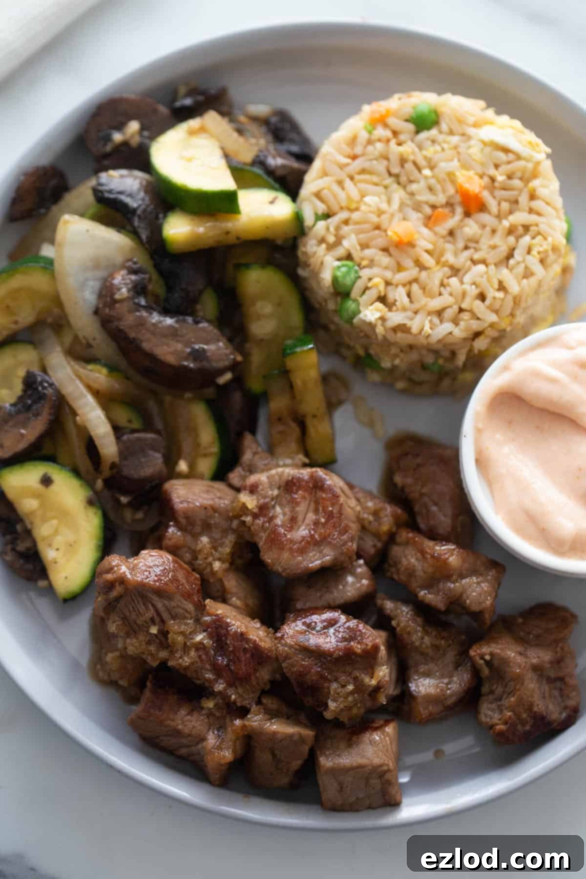 Sizzling Hibachi Steak 7 Overhead photo of white rimmed plate with hibachi steak cubes, sauteed veggies, fried rice, and a ramekin of yum yum sauce.