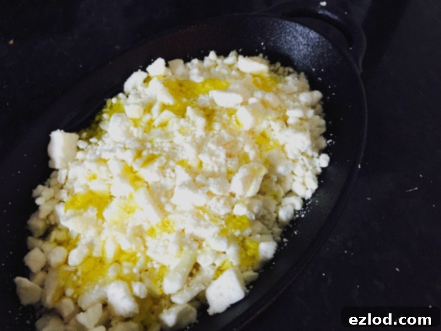 A cast iron pan with crumbled feta cheese drizzled with olive oil, ready for roasting, highlighting the simple preparation.
